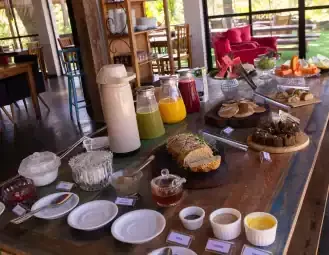 Imagem de um café da manhã com sucos, pães e doces em ambiente aconchegante.